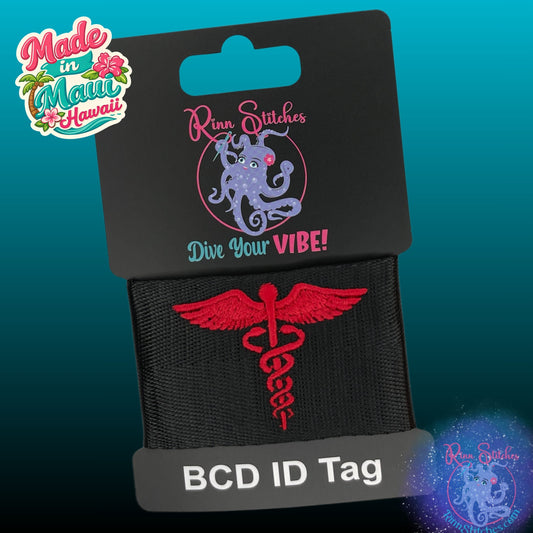 A black BCD ID tag featuring a red embroidered caduceus symbol, attached to a black display card with the brand name Rinn Stitches, made in Maui, Hawaii