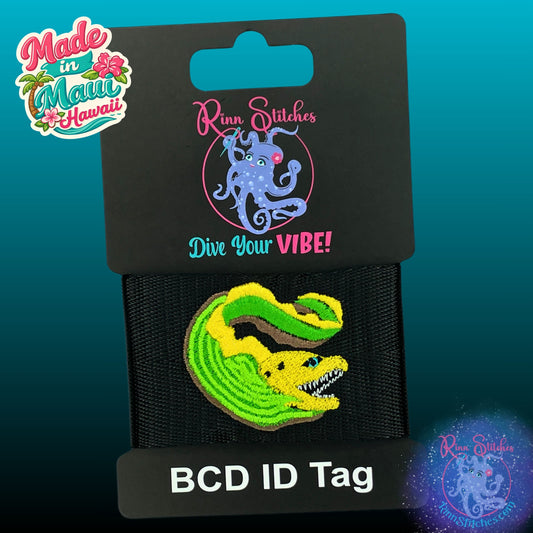 A black BCD ID tag featuring an embroidered green and yellow moray eel, presented on a black retail card with the brand name 'Rinn Stitches' and 'Dive Your VIBE!' printed on it. A 'Made in Maui Hawaii' logo is visible in the top left corner.