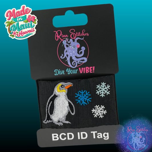 A BCD ID tag featuring an embroidered penguin and snowflakes on black webbing, attached to a black retail card with the brand name 'Rinn Stitches' and the phrase 'Dive Your VIBE!'.