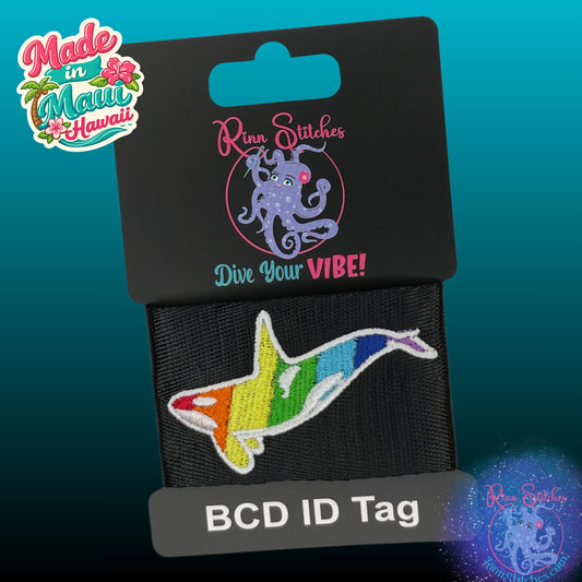 Rainbow-striped orca BCD ID tag on a black display card with the Rinn Stitches logo, shown against a teal gradient background. Made in Maui, Hawaii