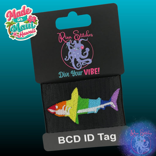 An embroidered BCD ID tag shaped like a rainbow-striped shark, attached to a black display card with the brand name 'Rinn Stitches' and 'Dive Your VIBE!' text. A 'Made in Maui Hawaii' logo is visible in the top left corner.