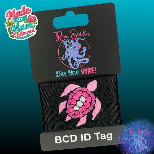 A BCD ID tag featuring a pink and white sea turtle design on a black webbing, displayed with its retail backing card that reads 'Rinn Stitches Dive Your VIBE!' and 'Made in Maui Hawaii'.