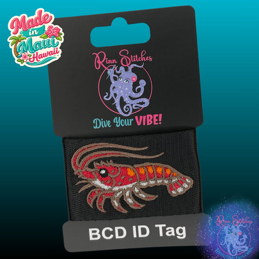 A Rinn Stitches BCD ID tag featuring an embroidered red, orange, and brown spiny lobster design on black webbing, attached to a black hanging card. Made in Maui, Hawaii