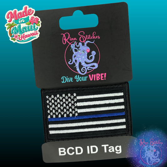 A BCD ID tag featuring a black and white American flag with a single thin blue line, attached to a black card display. Made by Rinn Stitches in Maui, Hawaii